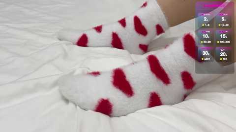 Video of a person lying on a white bed, wearing red heart-patterned white socks. A digital game overlay in the corner displays stats like \"2, 10, 15, 30, 20, 0, 0, 0.\