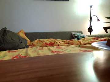 Video of a messy, unmade bed with a green blanket and yellow floral sheets, against a beige wall. A lamp with a white shade casts light on a wooden table.