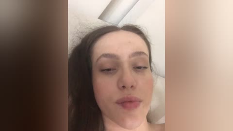 A close-up video of a young woman with light skin, long brown hair, and closed eyes, partially obscured by dark brown hair. The background is a white shower curtain with a metal rod.