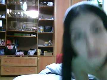 A blurry video of a person with short black hair, possibly a woman, standing in a cluttered room with wooden shelves filled with books and personal items, including a red kettle.