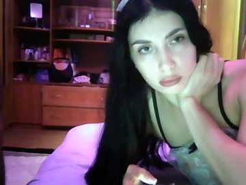 Video of a young woman with long black hair, wearing a black bra, lying on a bed with a wooden dresser and shelves in the background.