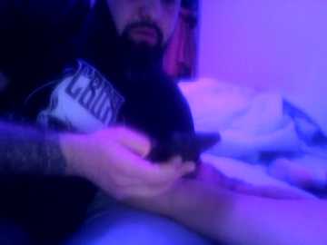 A blurry video with purple lighting shows a man with a beard, wearing a black hoodie, holding a small black object over a person's leg.