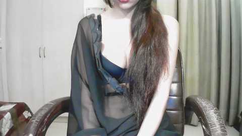 A video of a young woman with long, dark hair, wearing a sheer black robe that reveals a black bra, sitting on a brown leather chair in a dimly lit room with white walls and beige curtains.