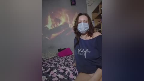 A candid video of a woman with a blue mask, off-shoulder dark sweater, and floral-patterned underwear, seated on a bed in a dimly lit room with a TV screen showing flames.