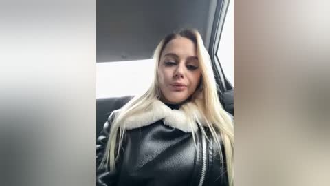 A video of a woman with long, straight blonde hair, wearing a black leather jacket with a white fur collar, seated in a car with grey upholstery.