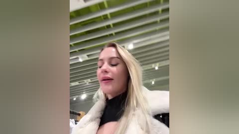Video of a fair-skinned, blonde woman with long hair, wearing a black top and white fur coat, standing indoors with green ceiling panels and modern lighting.