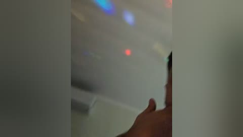 Video of a hand holding a white object, blurred background with colorful, abstract light patterns, suggesting a modern, minimalist interior.