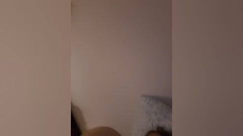 Video of a blurred, pale-skinned person's head and torso against a beige wall, with a hint of a bed or pillow in the background. The image is soft-focused and dimly lit, creating a sense of mystery and intimacy.