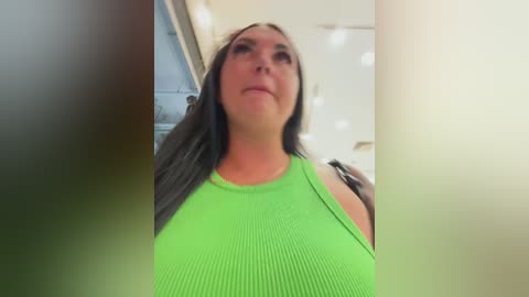 Video of a woman with long, straight brown hair, wearing a bright green, ribbed tank top, standing indoors with blurred background.