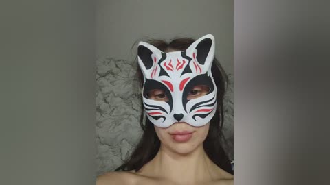 Video of a young woman with long dark hair, wearing a white, stylized fox mask with red and black designs, against a textured gray wall.