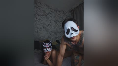 A video of a tattooed, shirtless man wearing a panda mask kneeling beside a woman in a cat mask on a bed in a dimly lit room with textured walls.