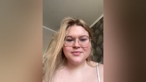 Video of a young, fair-skinned, plus-sized woman with long blonde hair, wearing glasses and a white tank top, smiling, in a room with beige walls and a stone-patterned backdrop.