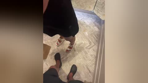 Video of a person wearing black shorts and red sandals standing on a beige and white tiled floor, viewed from above.