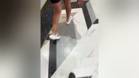 Video of a person in black shorts and white sneakers walking on a black and white geometric-patterned floor. The background is blurry, focusing on the person's legs.