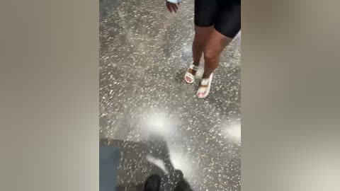 A video of a person's legs in black shorts and white sandals standing on a wet, gravelly surface. A dark shadow is cast on the ground.