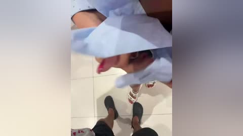 A video captures a woman in a light blue shirt, pink toenails, and gray slippers, standing in a room with a white tiled floor. A mirror reflects her face and upper body, partially obstructing her view.