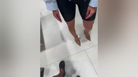 A video showing a person's legs and feet in black high heels reflected on a shiny white tiled floor, wearing a light blue shirt and black shorts.