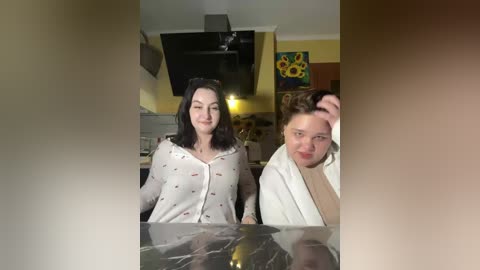 Video of two women in a cozy living room; one with straight black hair, the other with curly brown hair. They are dressed casually in white tops. The room features a TV, wall art, and a fireplace in the background.