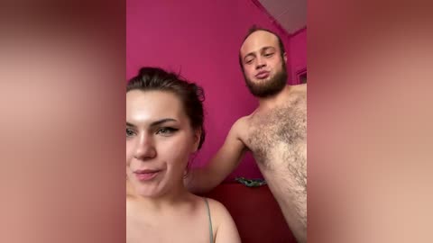 Video of a woman with dark hair and light skin, wearing a strapless top, standing beside a shirtless man with a beard, both against a bright pink wall.