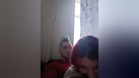 Video of two men, one with dyed blue hair and glasses, the other with red hair, in a dimly lit room with a window and white curtains.