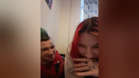 A video of two young people in a narrow, dimly-lit hallway. The person on the left has short green hair, a red shirt, and a bandaged nose, smiling. The person on the right has red hair, a white shirt, and tattoos on their fingers, looking down.