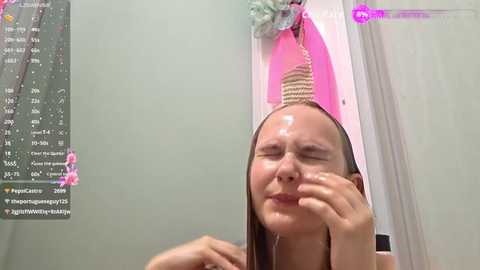 Video of a fair-skinned young woman with long brown hair, wearing a pink headband with a cat design, wiping her face with her right hand, in a bathroom with a calendar and pink decorations on the wall.