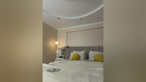 A video of a modern, elegant bedroom with beige walls, a grey upholstered headboard, a white bedspread, yellow pillows, a floral-patterned wallpaper, and a round white lamp on a side table.