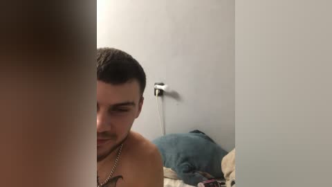 Video of a shirtless man with short dark hair, wearing a chain necklace, looking at his reflection in a mirror partially obscured by another person's arm. The background shows a messy bed with blue pillows and beige sheets.