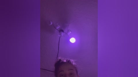 Video of a girl's face with a light purple glow, looking upwards at a cracked ceiling light bulb emitting a bright white light.