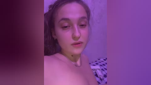 A video of a young woman with light skin and shoulder-length brown hair, wearing a necklace, in a dimly lit room with purple lighting.