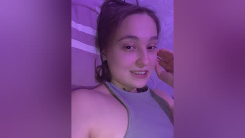 A young woman with fair skin, brown hair, and light makeup, wearing a gray top, smiles while touching her cheek in a dimly lit room.