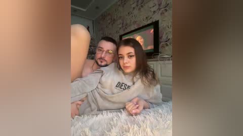 Video of a man and woman, both with light skin, lying on a soft, white shag rug in a cozy, floral-patterned room. The man, with glasses, wears a grey sweatshirt, while the woman, with long brown hair, wears a grey sweatshirt.