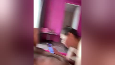 Blurry video of a woman with dark hair, possibly East Asian, sitting on a bed in a pink room, holding a phone. The image is slightly out of focus.