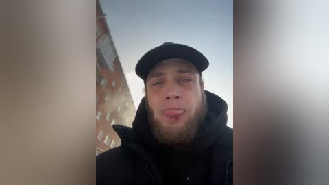 Video of a young man with fair skin, a beard, and a black cap, sticking out his tongue, taken from a low angle, with a red brick building in the background.