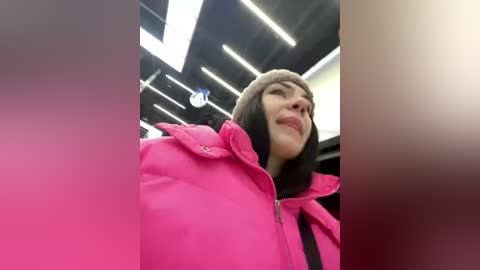 Video of a young woman with light skin, brown hair, and a pink jacket, smiling inside a brightly lit, modern indoor space with white fluorescent lights and a blurred background.