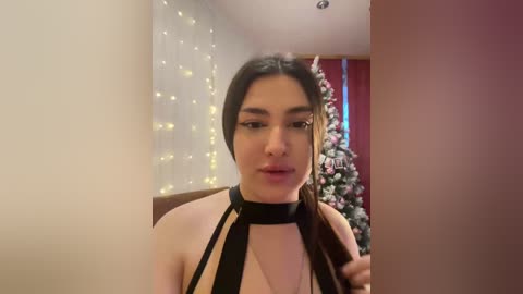A video of a young woman with light skin and dark hair, wearing a black halter top, standing in a festive room with string lights and a decorated Christmas tree.