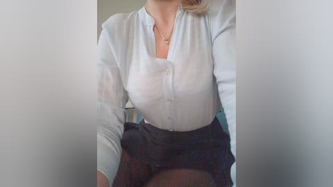 Video of a woman with fair skin and blonde hair, wearing a white blouse and a dark skirt, sitting indoors with blurred background.