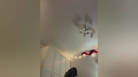 A video of a ceiling fan with a white light fixture, and a red ribbon draped across the room, with white cabinets and a dark object partially visible.