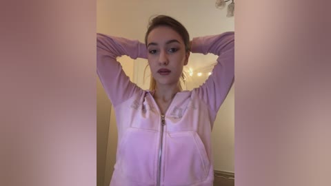 Video of a young Caucasian woman with fair skin, light brown hair tied back, wearing a pink hoodie, stretching her arms behind her head in a cozy indoor setting with a white door and a blurred background.