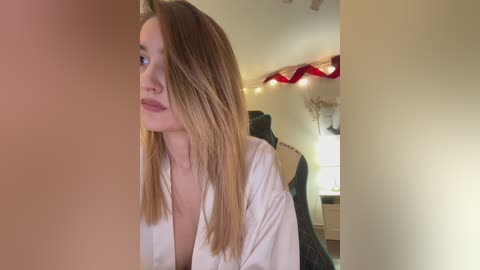 Video of a fair-skinned woman with long, straight blonde hair, wearing a white robe, looking pensive in a dimly lit bedroom with a green chair and string lights in the background.