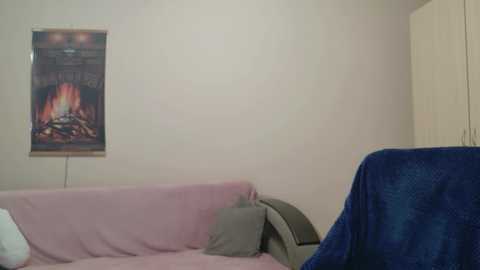 A video of a cozy, minimalistic living room featuring a pink sofa draped with a blue blanket, a wall-mounted fireplace with a burning fire, and a white wall.