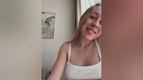 Video of a smiling Caucasian woman with long blonde hair, wearing a white ribbed tank top, standing in a minimalist room with beige walls and abstract art.