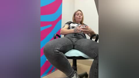 Video of a blonde woman in grey jeans and a black t-shirt, sitting on a chair, holding a phone, against a vibrant blue and pink wall in a modern room.