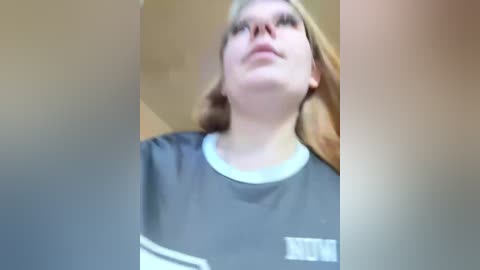 A blurred, close-up video of a young woman with fair skin, long blonde hair, and glasses, wearing a dark gray T-shirt. The background is indistinct.