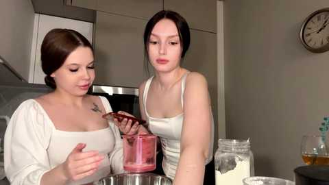 Video of two young women in a modern kitchen. One is pouring red sauce from a jar into a blender, while the other observes. Background includes stainless steel appliances, a clock, and a glass jar of milk.