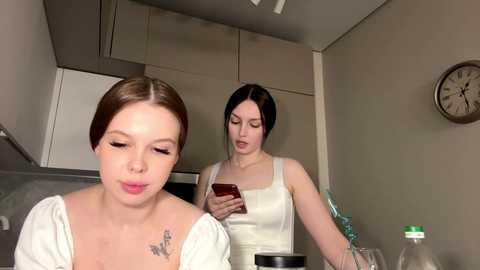 Video of a modern kitchen with two women: a brunette in a white shirt, a blonde in a white shirt, both looking down at a smartphone.
