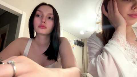 Video of a young Caucasian woman with straight black hair, wearing a white lace top, sitting in a dimly lit bedroom with cream walls and a ceiling light.