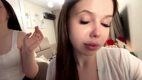 Video of a young woman with long brown hair, wearing a white top, applying makeup, in a cozy living room with beige furniture and modern decor.