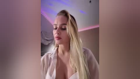 Video of a fair-skinned, blonde woman in a white shirt, standing in a dimly lit room with purple LED lights. Her expression is serene, and her hair is loose.
