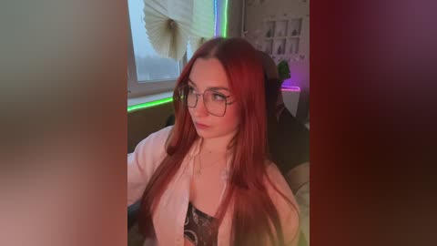 Video of a young, fair-skinned woman with long, straight red hair, wearing glasses and a white blazer over a black top, sitting indoors with neon lights and a sun-shaped poster in the background.